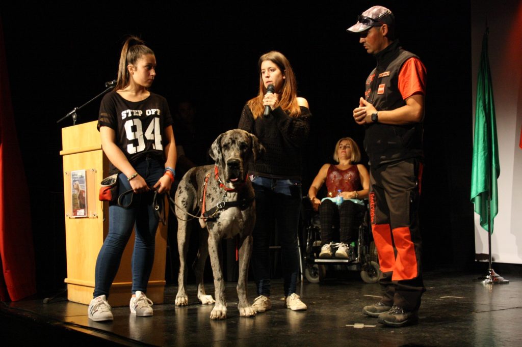Personas en un escenario junto a un perro grande con arnés; una mujer sostiene la correa y otra habla por un micrófono.