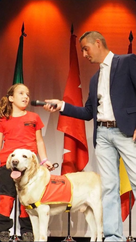 Hombre con micrófono frente a una niña y un perro claro con chaleco rojo, sobre un escenario.